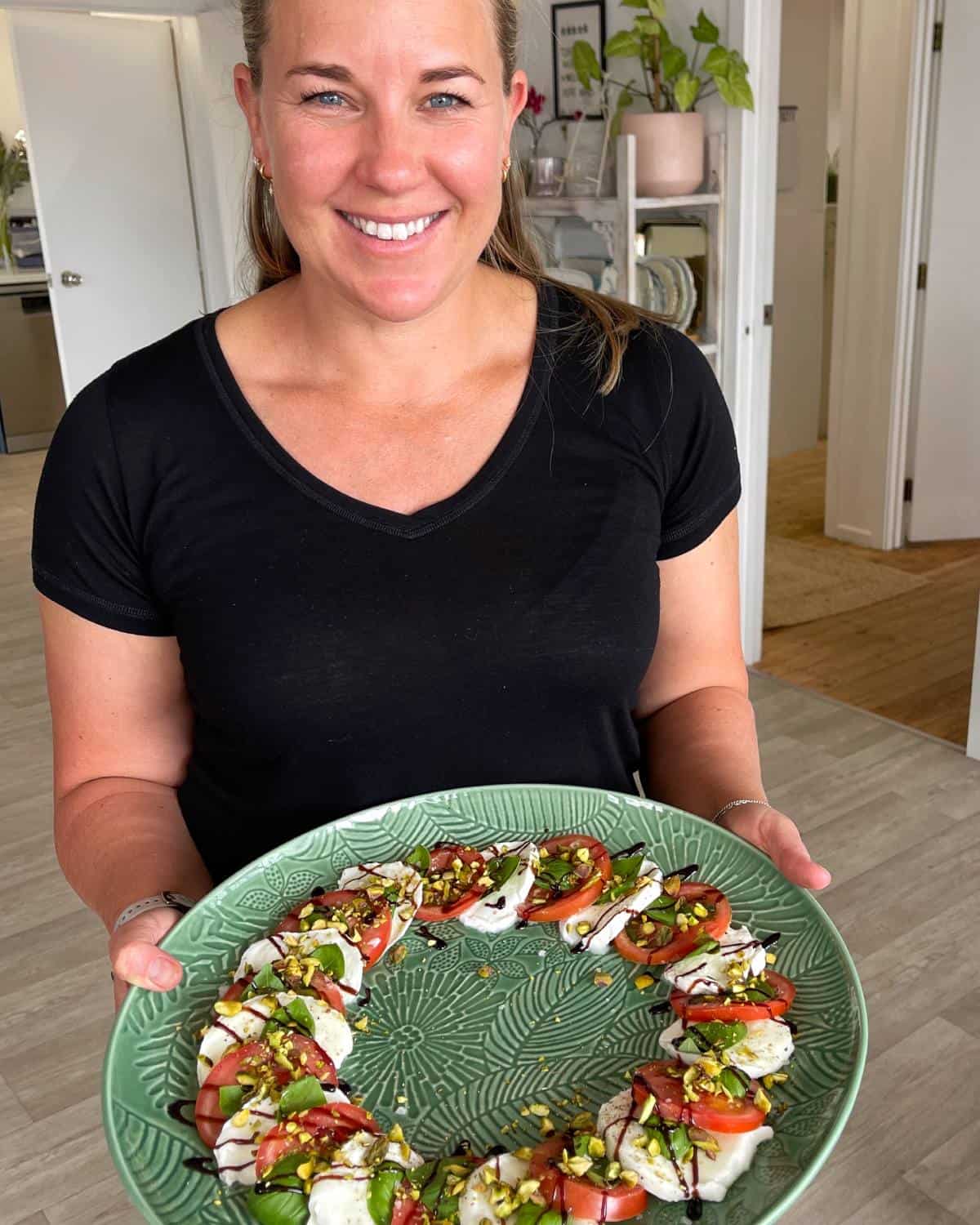 Caprese Wreath A woman in a black shirt holds a decorative green plate with a neatly arranged caprese salad topped with balsamic glaze and chopped nuts.