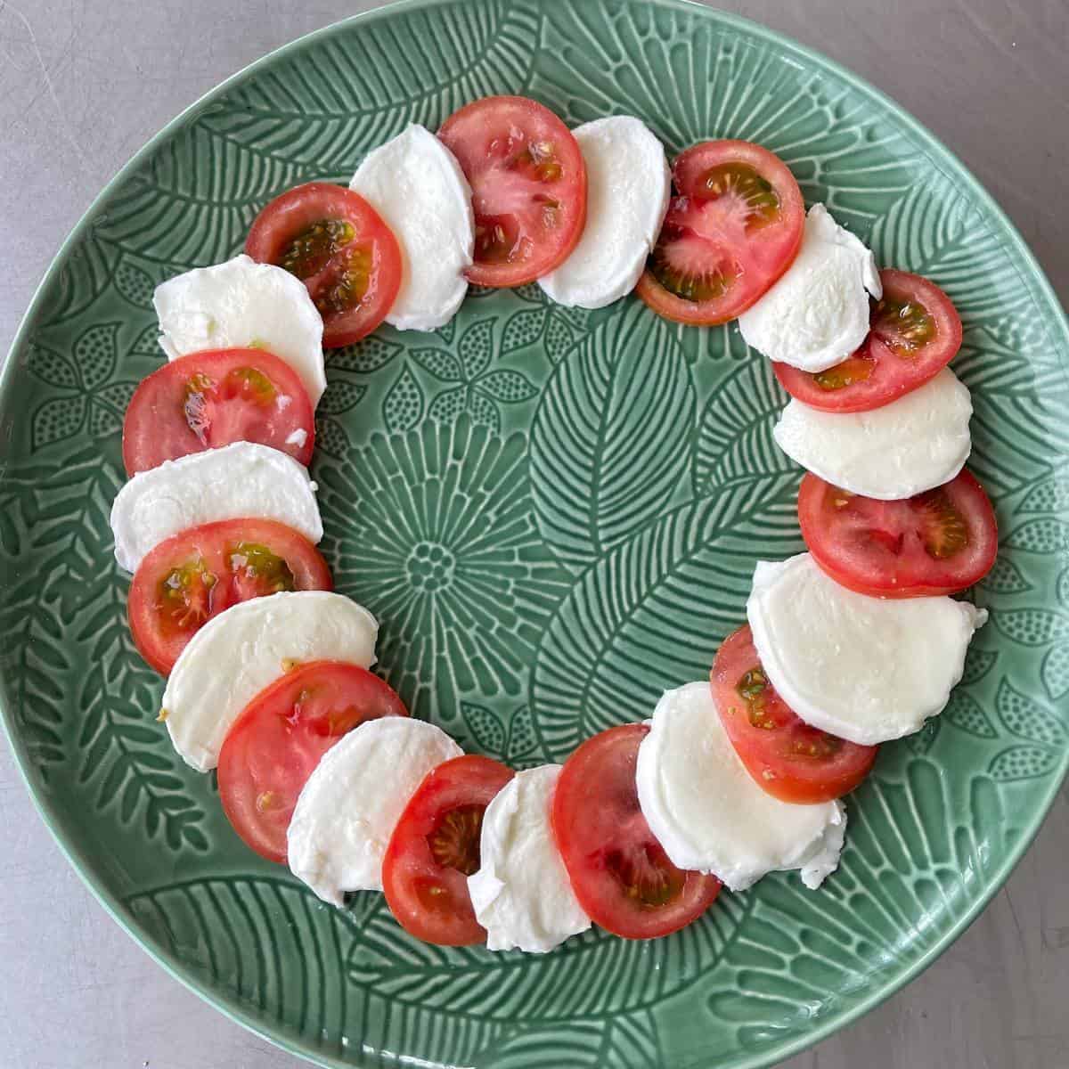 Caprese Wreath Tomato and mozzarella slices arranged alternately in a circular pattern on a decorative green plate.