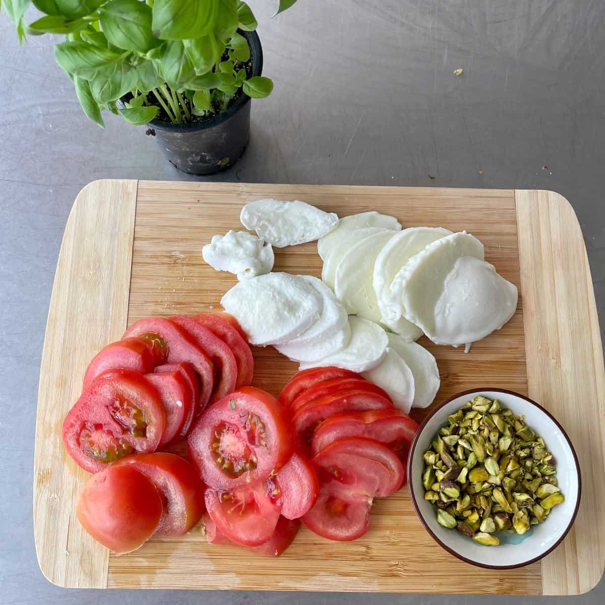 Caprese Wreath Sliced tomatoes, mozzarella cheese, and chopped pistachios are arranged on a wooden cutting board next to a small potted basil plant.