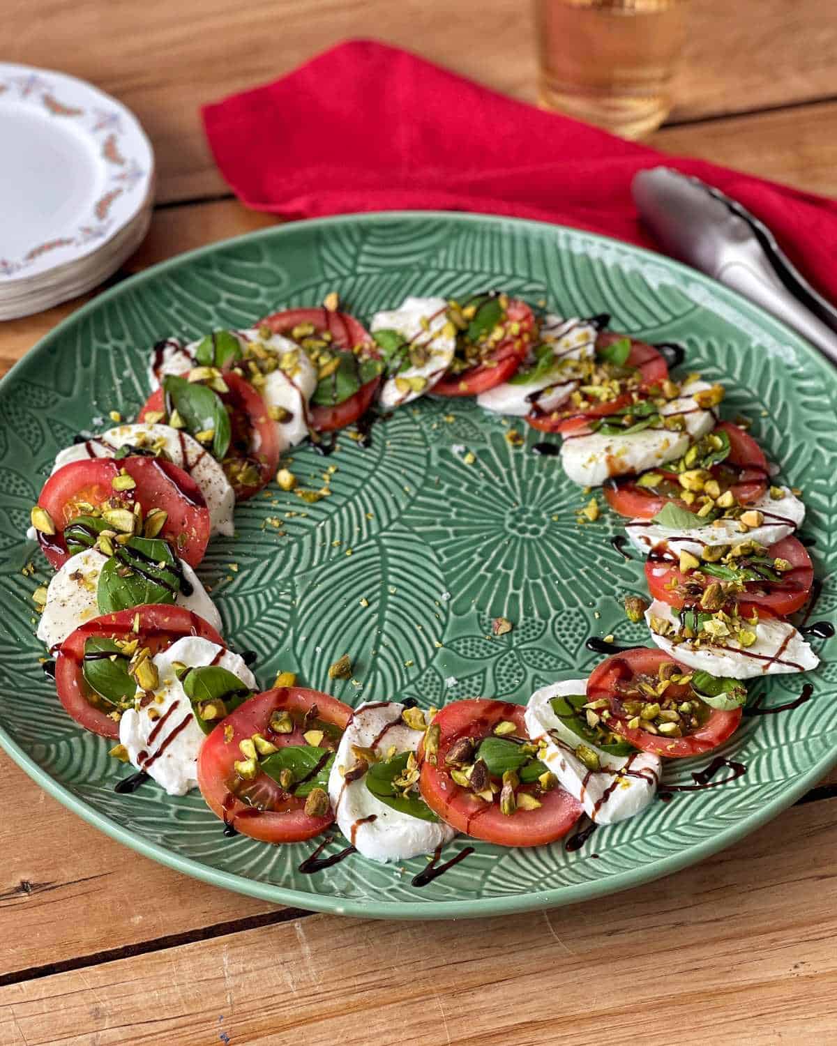 A plate of caprese salad arranged in a ring, featuring sliced tomatoes, mozzarella, basil, chopped pistachios, and a drizzle of balsamic glaze on a green patterned platter.