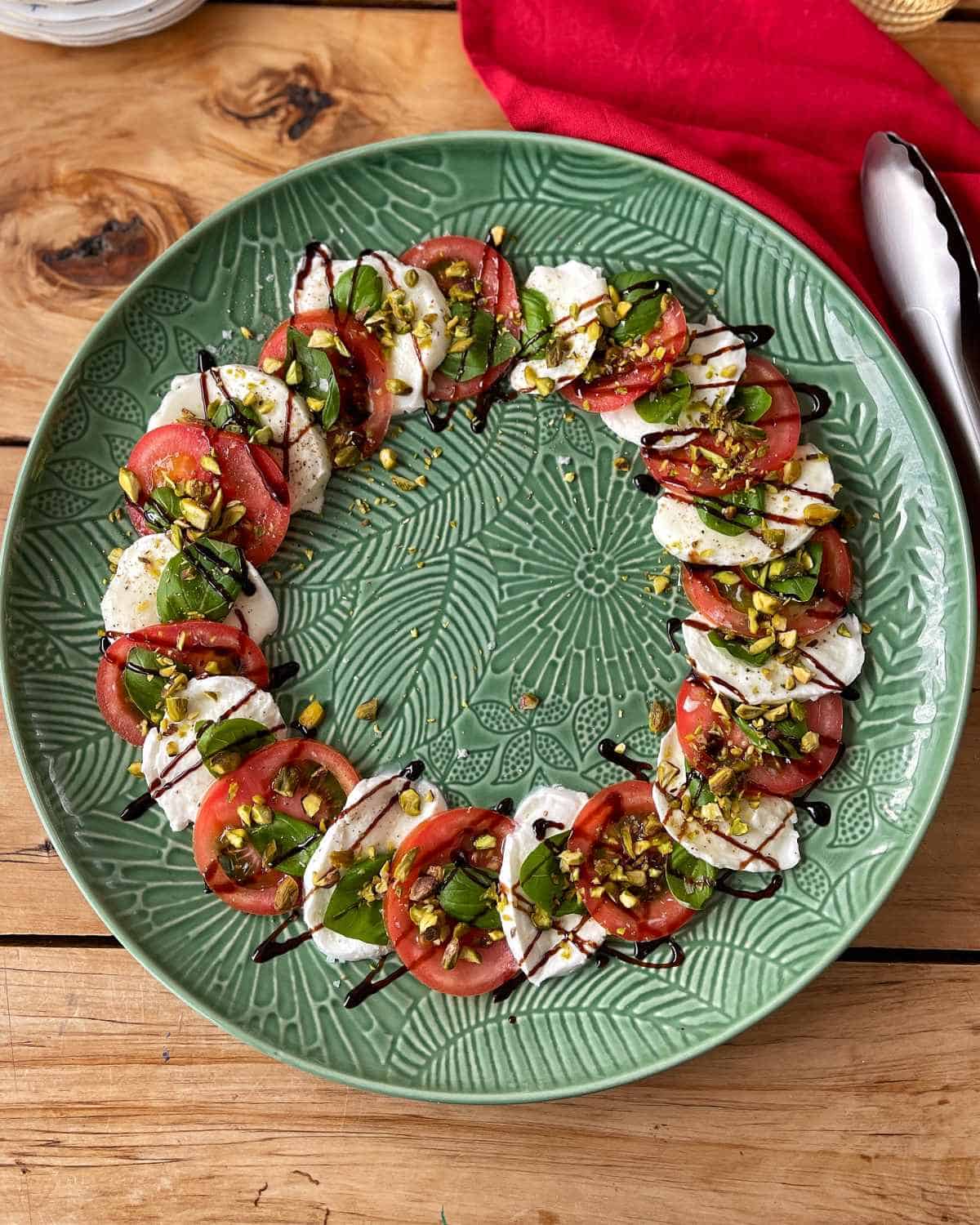 Caprese Wreath A circular arrangement of sliced tomatoes, mozzarella, and basil on a green patterned plate, drizzled with balsamic glaze and sprinkled with chopped nuts.