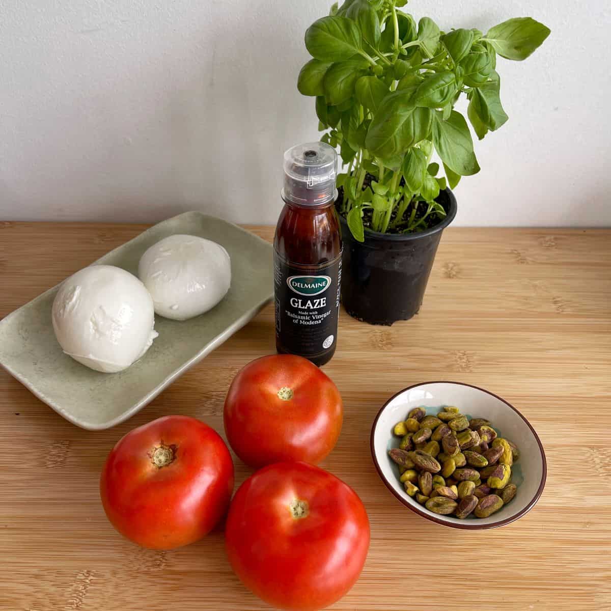 Caprese Wreath Mozzarella balls, fresh basil plant, balsamic glaze bottle, three tomatoes, and a bowl of pistachios arranged on a wooden surface.