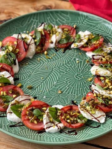 Sliced tomatoes, mozzarella, and basil arranged in a circle on a green plate, topped with chopped nuts and balsamic glaze.