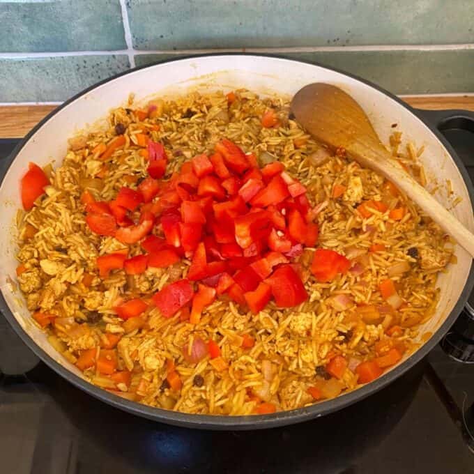 Chicken and Rice Pilaf A pan of rice mixed with vegetables and chicken, topped with freshly chopped red bell peppers, sits on a stovetop with a wooden spoon resting inside.