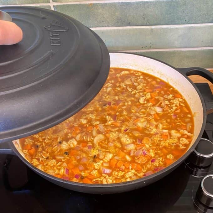 Chicken and Rice Pilaf A hand lifts the lid of a black pot revealing a simmering mixture of chopped vegetables and ground meat in a tomato-based sauce on a stovetop.