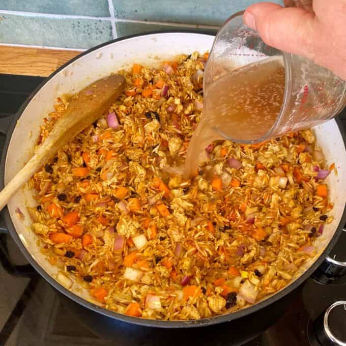 Chicken and Rice Pilaf A hand pours broth from a measuring cup into a pan of rice, chopped vegetables, and seasonings on a stovetop, with a wooden spoon resting in the pan.