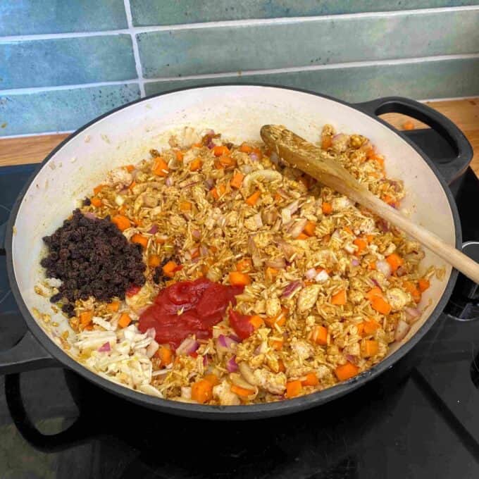 Chicken and Rice Pilaf A pot on a stove contains chopped vegetables, tomato paste, currants, and spices being cooked and mixed with a wooden spoon.