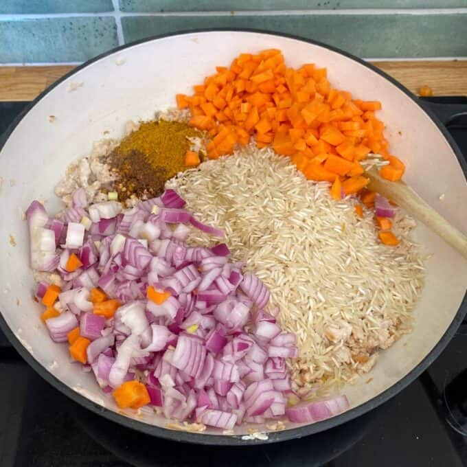 Chicken and Rice Pilaf A white pan on a stovetop contains uncooked rice, diced red onion, chopped carrots, minced meat, and curry powder, with a wooden spoon on the side.
