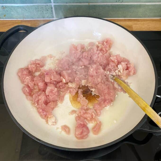 Chicken and Rice Pilaf Raw minced meat is being cooked in a white pan on a stovetop, with a wooden spoon stirring the mixture. Steam is rising from the pan.