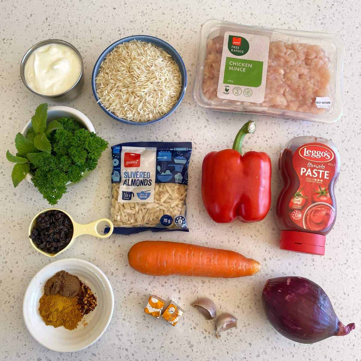 Chicken and Rice Pilaf A selection of ingredients on a countertop, including chicken mince, rice, red pepper, carrot, herbs, yogurt, slivered almonds, tomato paste, onion, garlic, spices, raisins, and stock cubes.