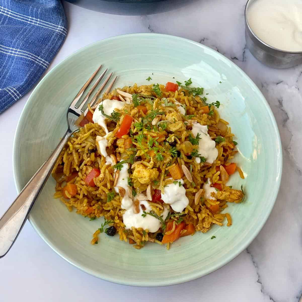 Chicken and Rice Pilaf A plate of spiced rice with vegetables and chicken, topped with a white yogurt sauce, with a fork on the side. A small bowl of sauce and a blue napkin are nearby.