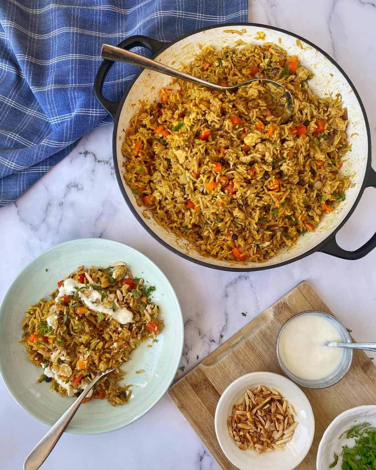 A pan of vegetable rice with a serving on a plate, garnished with sauce, next to bowls of yogurt and sliced almonds on a marble surface with a blue towel.