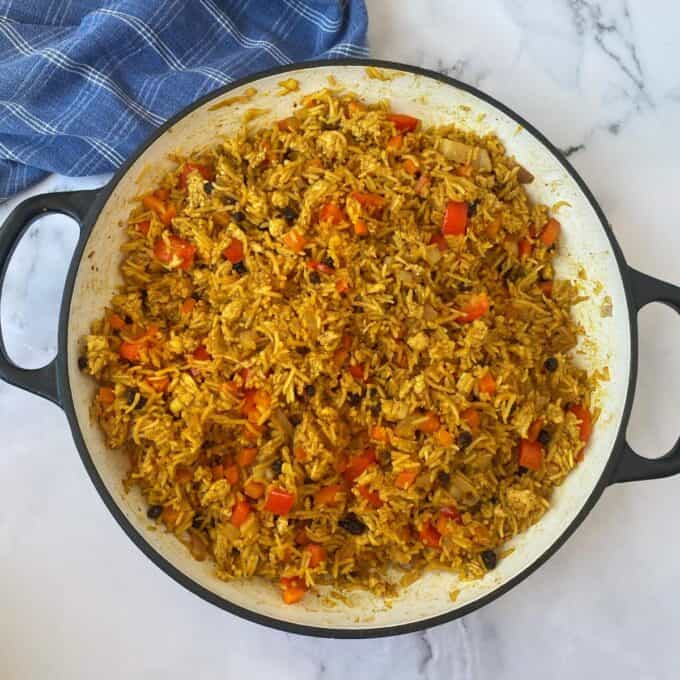 Chicken and Rice Pilaf A large white skillet contains cooked yellow rice mixed with diced red bell peppers, onions, and small dark bits, resting on a marble surface next to a blue cloth.
