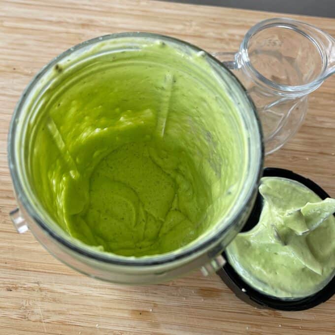 Green Goddess Avocado Salad A blender cup filled with thick green sauce sits on a wooden surface next to its lid, which also has some sauce on it, and an empty glass pitcher.