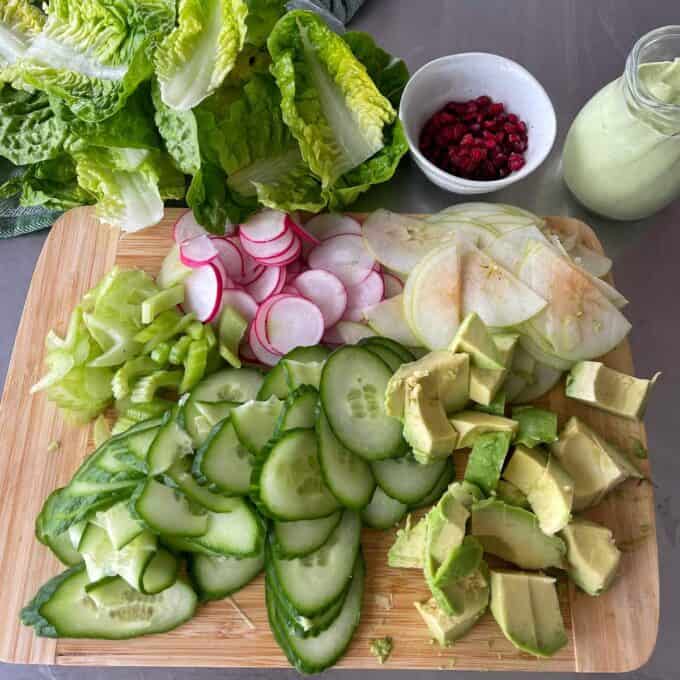 Green Goddess Avocado Salad A wooden board with sliced cucumber, avocado, radish, celery, and apple; nearby are romaine lettuce, a jar of dressing, and a small bowl of pomegranate seeds.