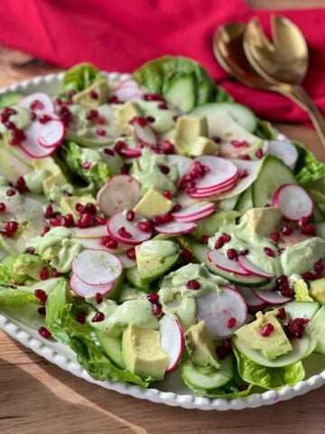 A salad with lettuce, sliced cucumber, radish, avocado chunks, and pomegranate seeds topped with a creamy green dressing on a white platter. Gold utensils and a red napkin are nearby.