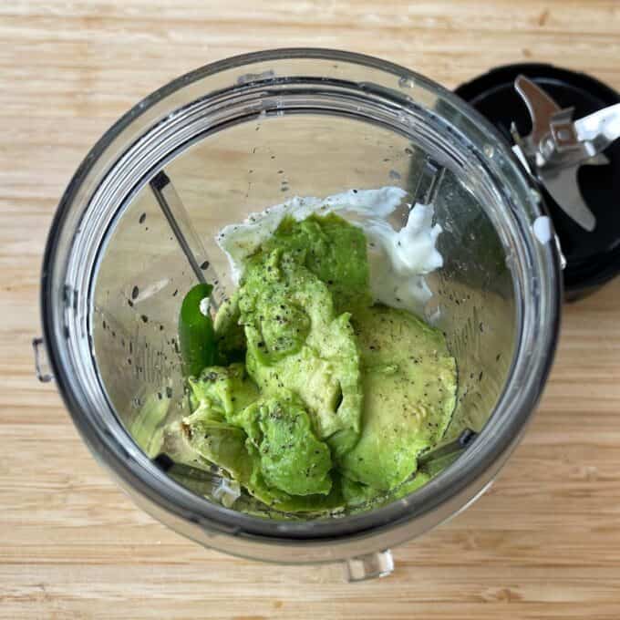 Green Goddess Avocado Salad A blender cup containing avocado, yogurt, and a piece of jalapeño, with a detached blade lid on the side, set on a wooden surface.