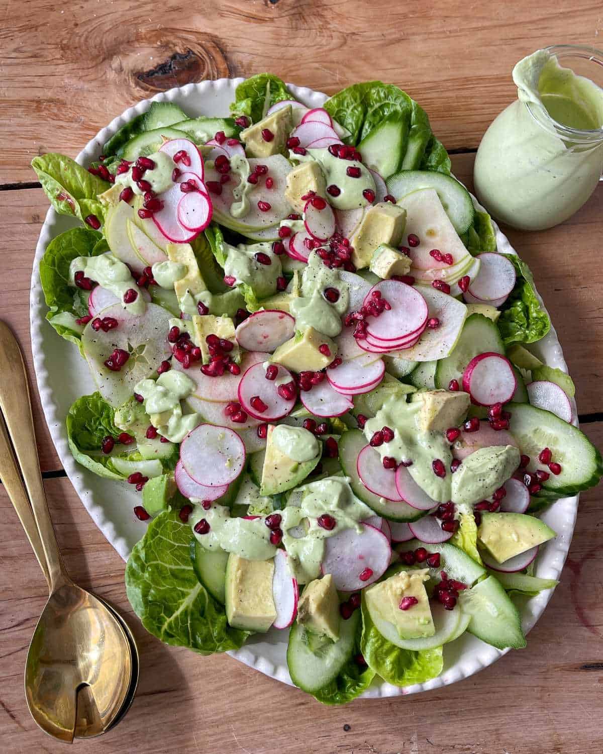 Green Goddess Avocado Salad Salad with romaine lettuce, sliced cucumber, radish, avocado, and pomegranate seeds, drizzled with a creamy green dressing, served on a white oval plate with gold utensils.