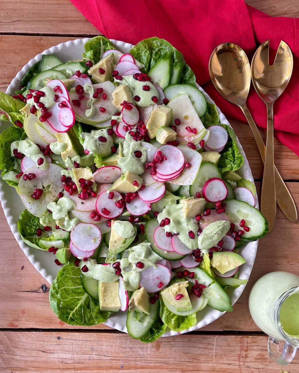 A platter of salad with lettuce, cucumber, radish, avocado, and pomegranate seeds, drizzled with green dressing. Gold serving utensils and a red napkin are beside it.