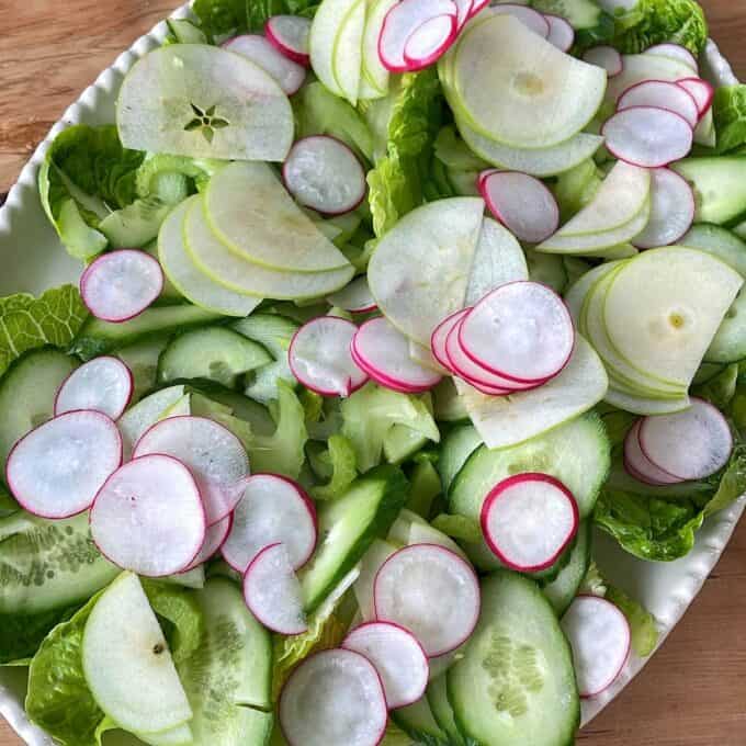 Green Goddess Avocado Salad A platter of fresh salad with sliced cucumbers, radishes, green apples, and leafy greens arranged on a white dish.