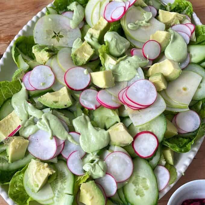 Green Goddess Avocado Salad A salad with sliced cucumbers, radishes, green apples, avocado chunks, and green dressing on top, served on a white plate.