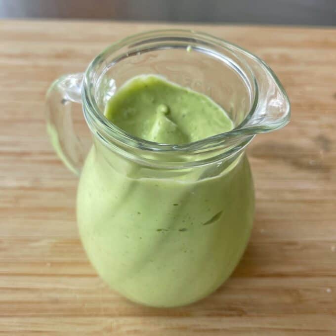 Green Goddess Avocado Salad A glass pitcher filled with creamy green sauce sits on a wooden surface.