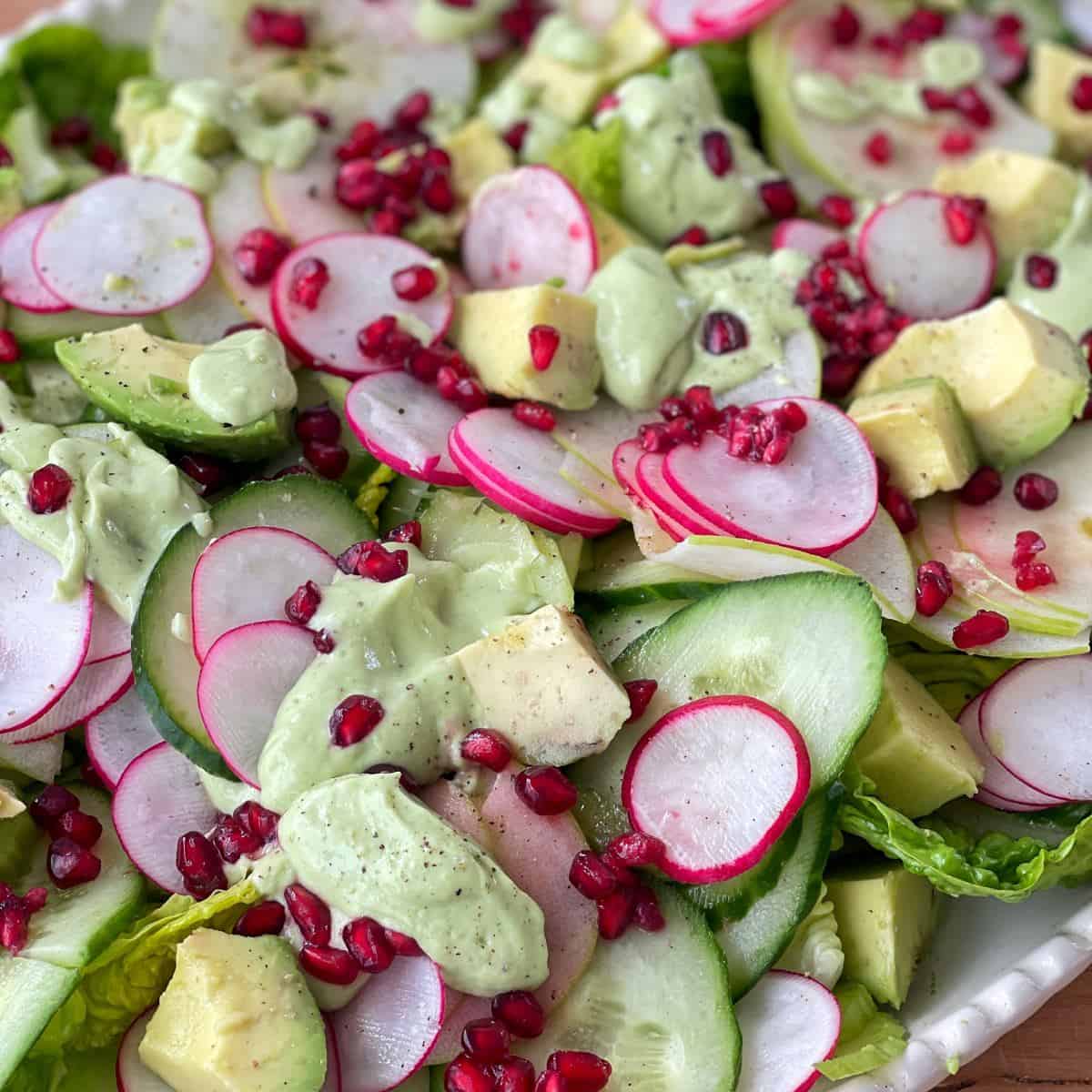 Green Goddess Avocado Salad A fresh salad with sliced cucumbers, radishes, avocado chunks, pomegranate seeds, and green dressing on a bed of leafy greens.