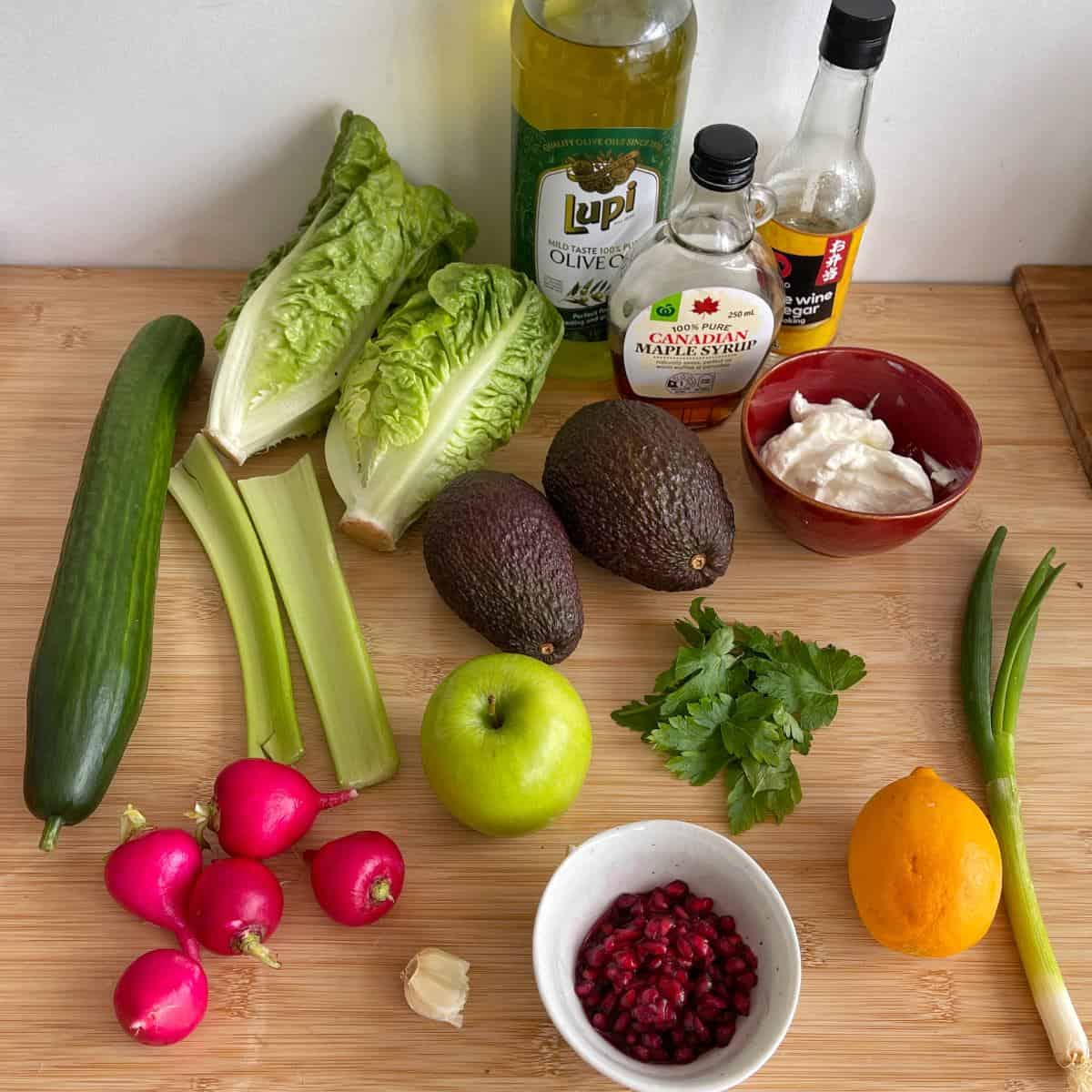 Green Goddess Avocado Salad Various fresh ingredients including lettuce, celery, cucumber, radishes, apple, avocado, lemon, parsley, green onion, pomegranate seeds, garlic, mayonnaise, olive oil, maple syrup, and soy sauce.