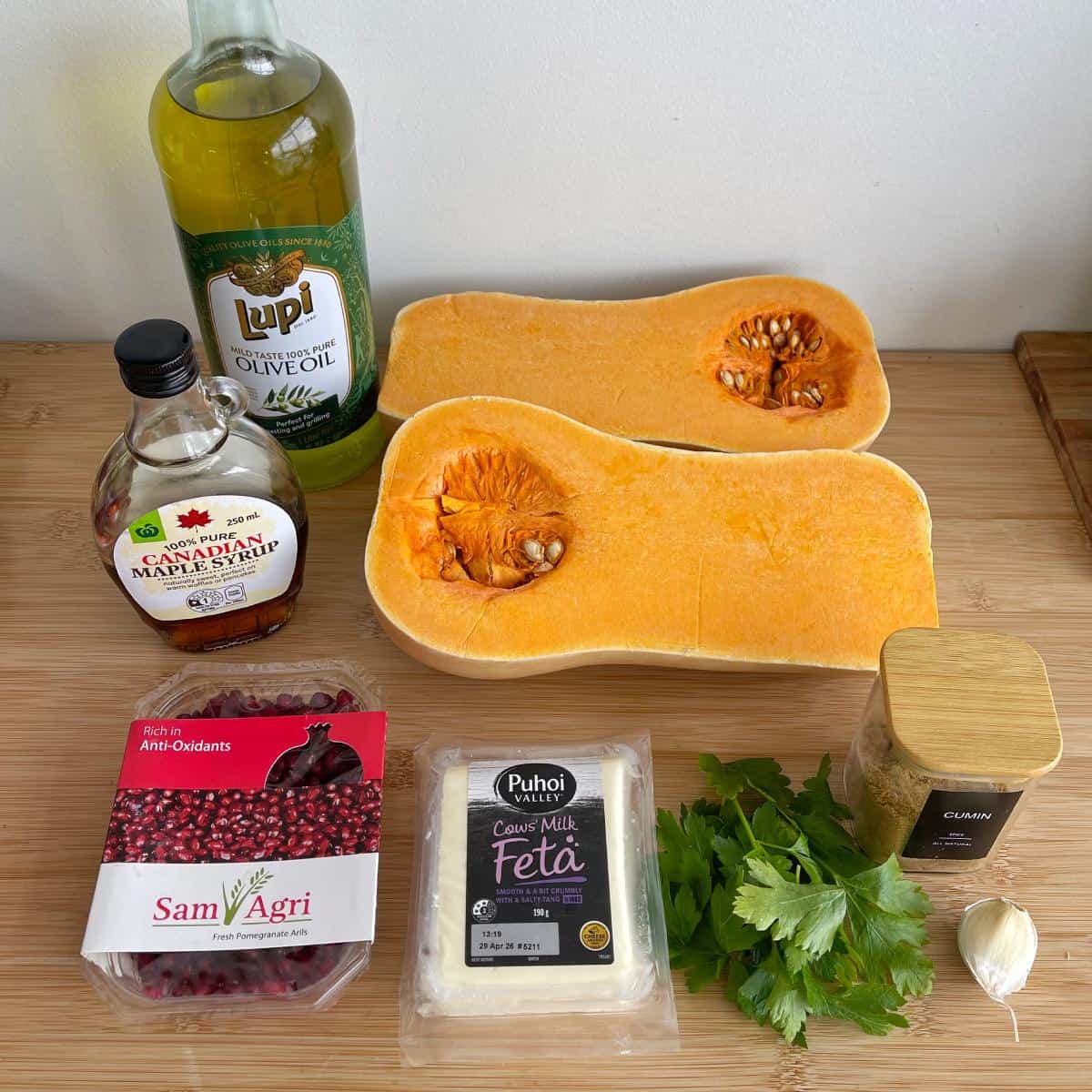Maple Roasted Hasselback Butternut A bottle of olive oil, halved butternut squash, maple syrup, cumin, garlic, flat-leaf parsley, feta cheese, and pomegranate seeds on a wooden surface.