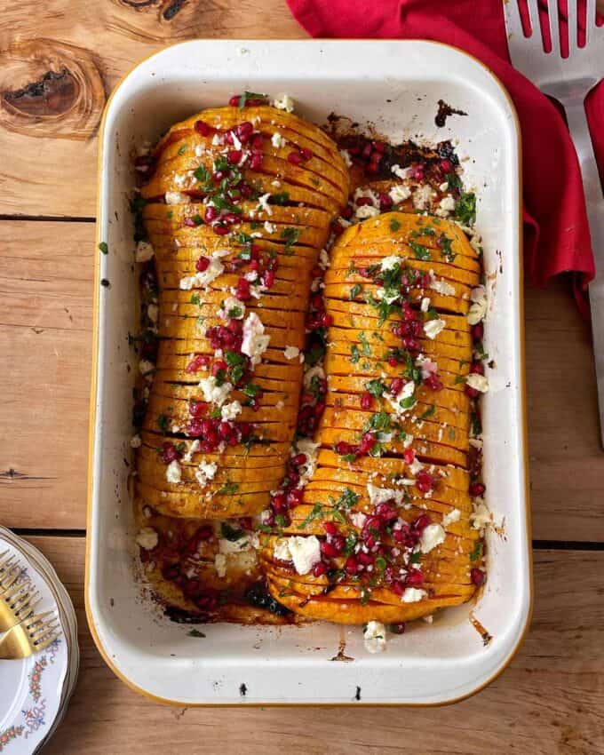 Maple Roasted Hasselback Butternut Two roasted hasselback butternut squash halves topped with feta, pomegranate seeds, and herbs in a white baking dish on a wooden table.