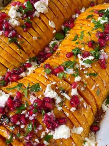 Sliced roasted butternut squash in a baking dish, topped with crumbled cheese, pomegranate seeds, and chopped herbs.