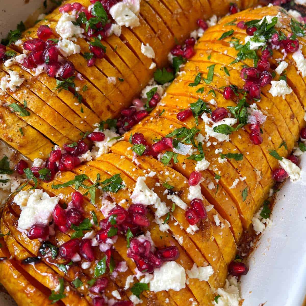 Maple Roasted Hasselback Butternut Sliced roasted butternut squash in a baking dish, topped with crumbled cheese, pomegranate seeds, and chopped herbs.