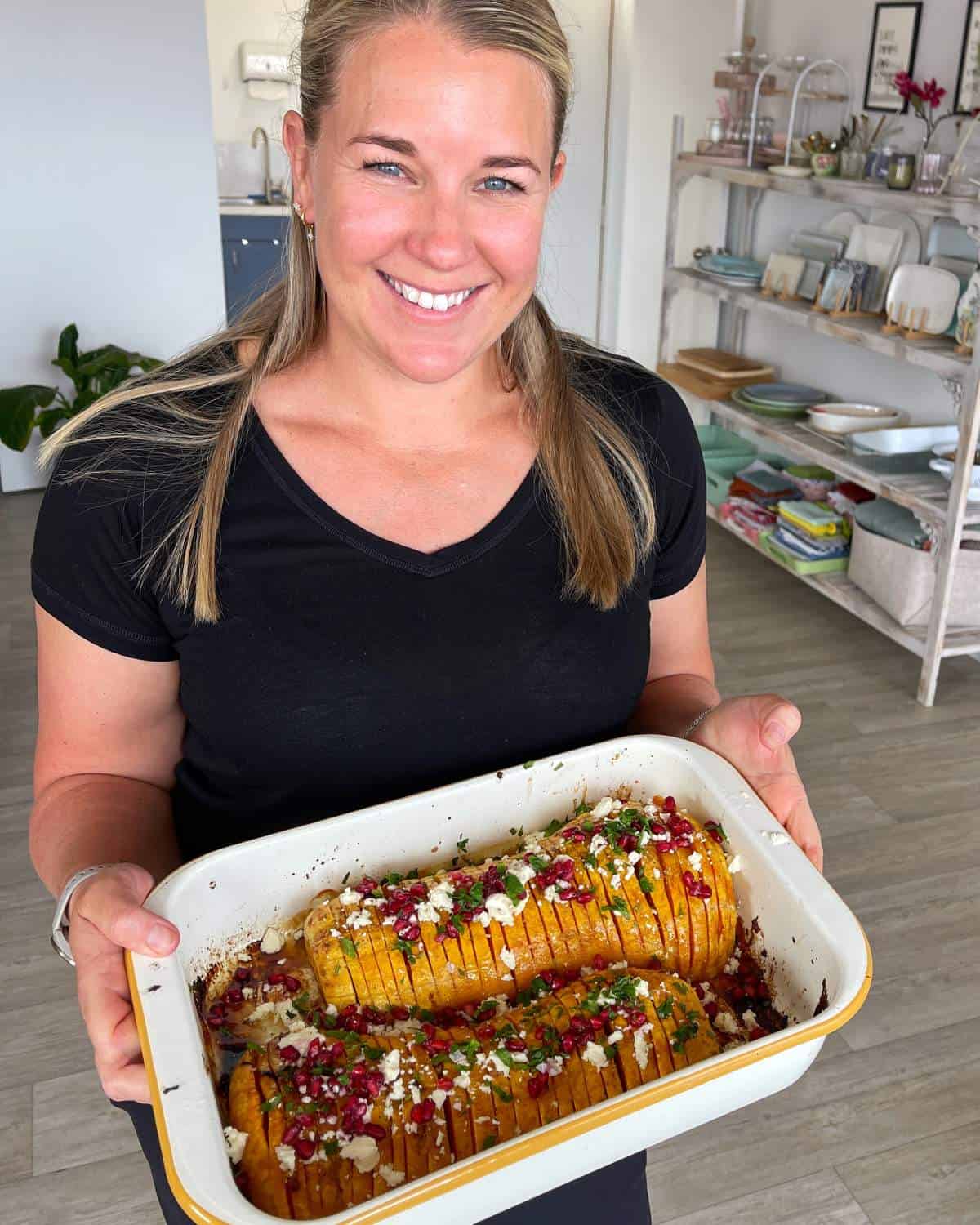 Maple Roasted Hasselback Butternut A woman in a black shirt smiles while holding a baking dish with two cooked, sliced squashes topped with herbs and cheese in a kitchen setting.