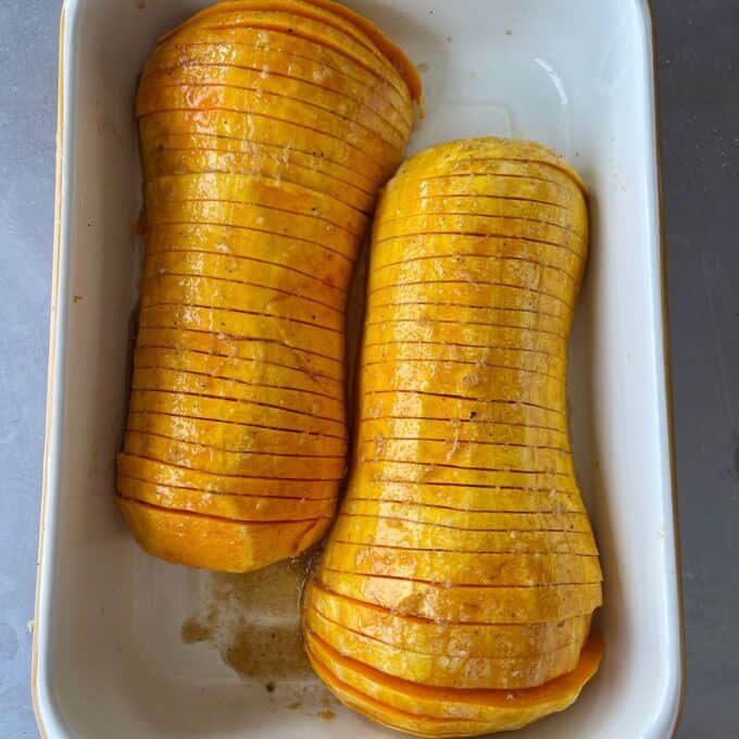 Maple Roasted Hasselback Butternut Two hasselback butternut squashes are sliced and arranged side by side in a white baking dish, with some melted butter at the bottom.