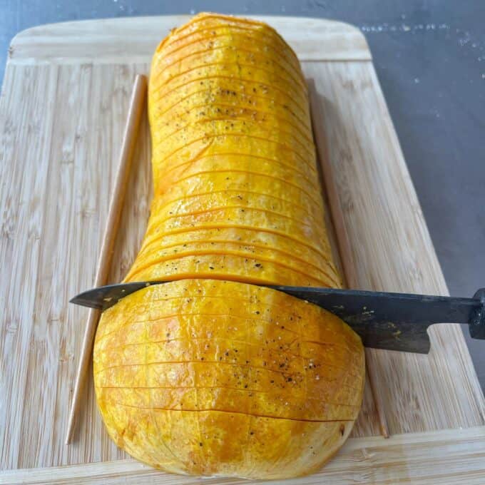 Maple Roasted Hasselback Butternut A roasted butternut squash is being sliced with a knife on a wooden cutting board.