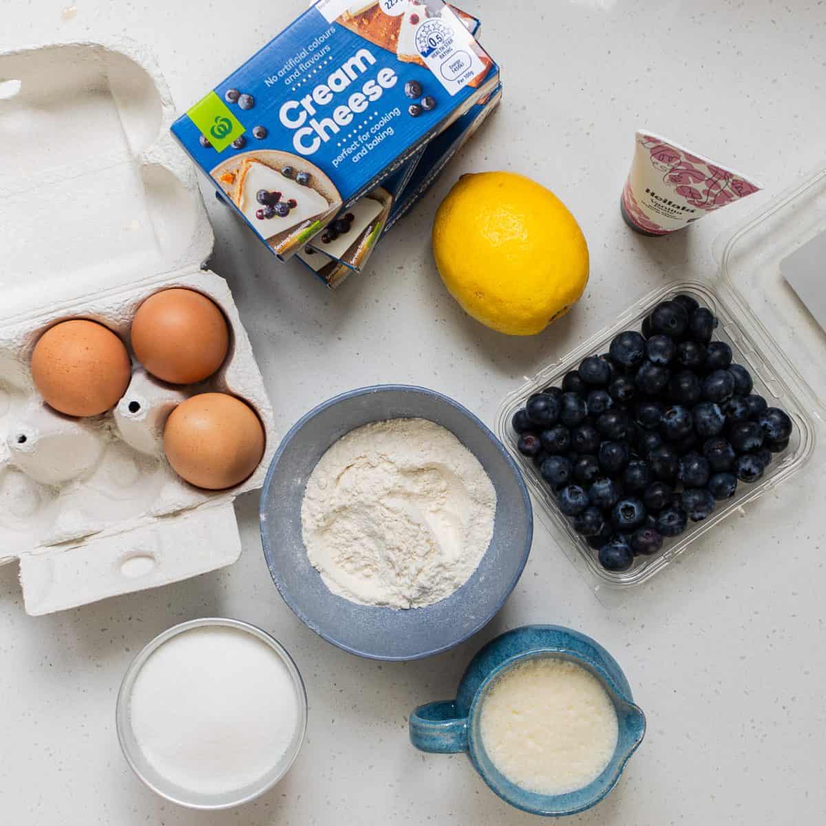 Lemon Blueberry Basque Cheesecake Ingredients on a counter: three eggs, cream cheese packages, a lemon, blueberries, a bowl of flour, a cup of milk, a bowl of sugar, and a small carton of cream.