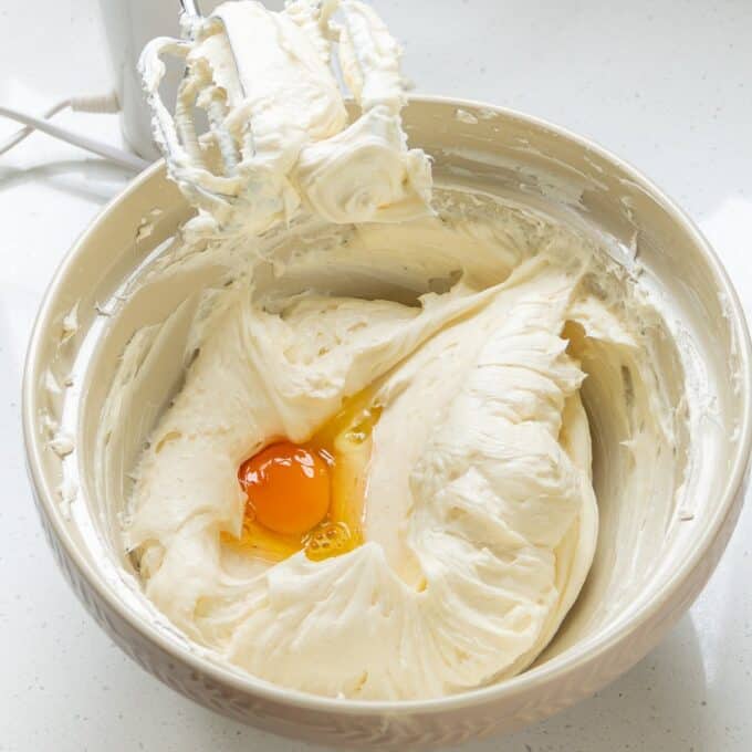 Lemon Blueberry Basque Cheesecake A mixing bowl containing creamy batter with an egg added on top, next to a mixer with beaters covered in batter.