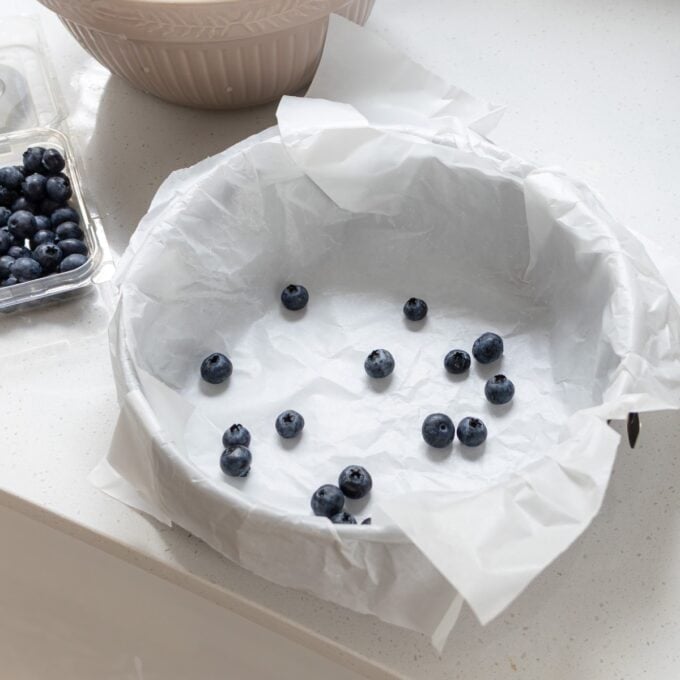 Lemon Blueberry Basque Cheesecake Round baking pan lined with parchment paper, scattered with a few blueberries, sitting on a white countertop next to a plastic container of more blueberries and a mixing bowl.