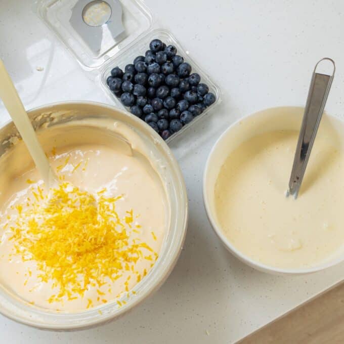 Lemon Blueberry Basque Cheesecake A bowl of batter with lemon zest, a bowl with a whisk and batter, and a container of blueberries on a countertop.