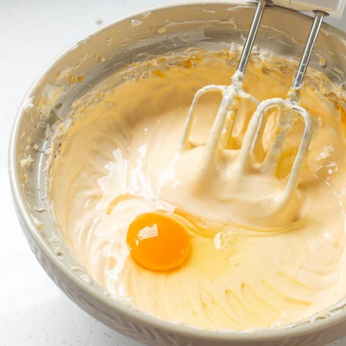 Lemon Blueberry Basque Cheesecake Electric mixer beaters in a bowl of creamy batter with a raw egg added on top, ready to be mixed in.