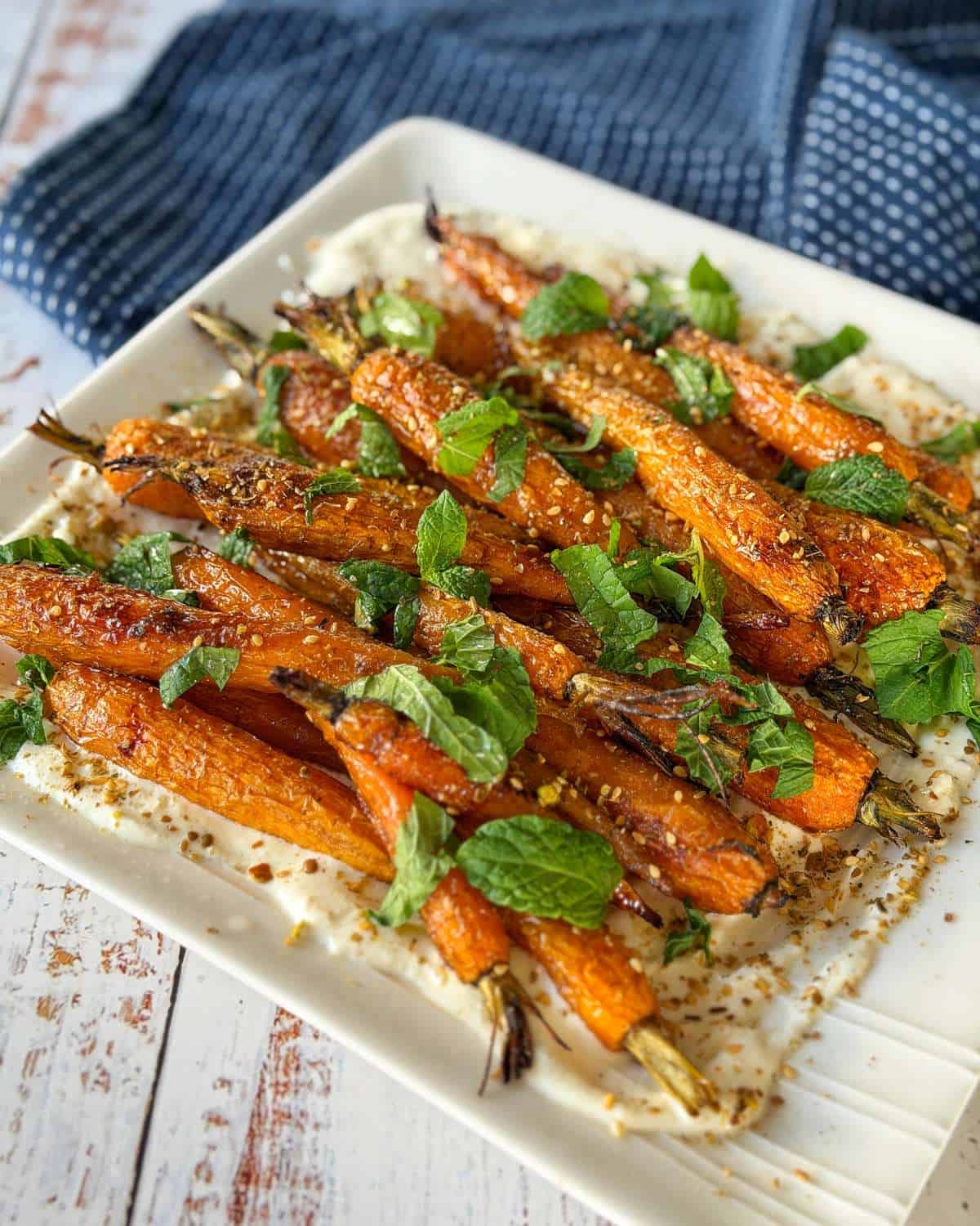 Roasted carrots garnished with fresh mint leaves and spices, served on a white square plate with a blue cloth in the background.