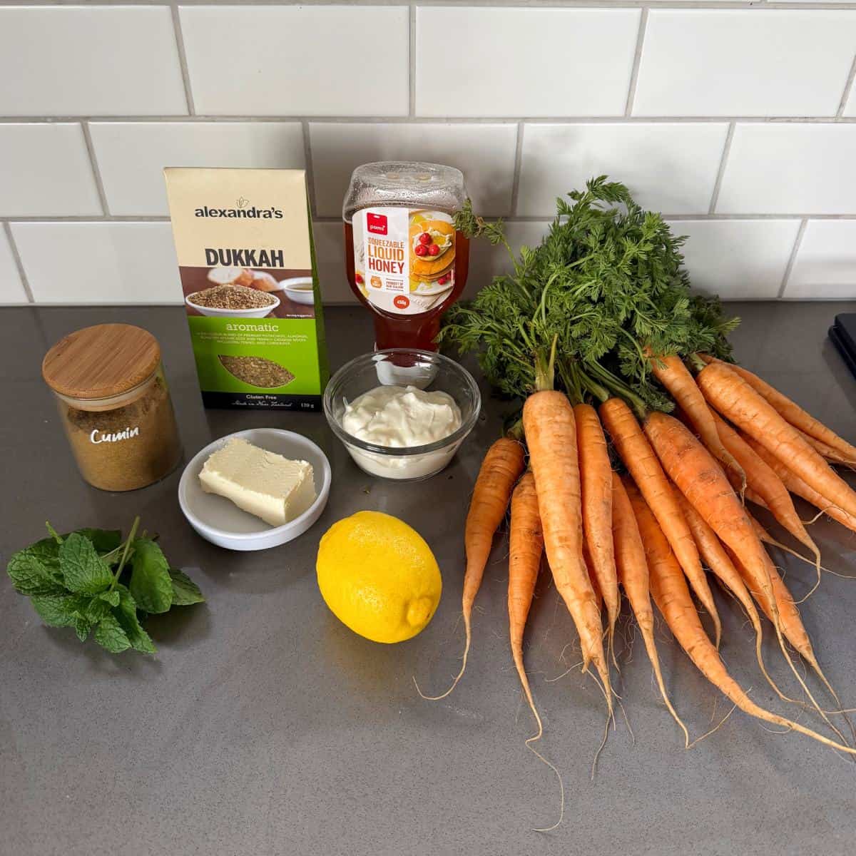 Roasted Carrot with Feta Whip A bunch of carrots with tops, a lemon, fresh mint, cumin powder, feta cheese, Greek yogurt, honey, and dukkah are arranged on a kitchen counter.