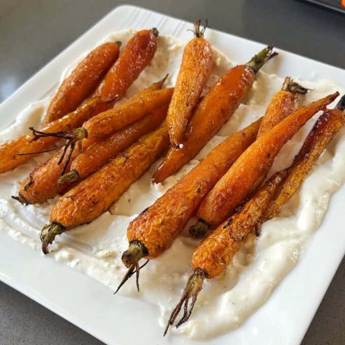 Roasted Carrot with Feta Whip A square white plate with roasted carrots arranged on a layer of creamy white sauce.
