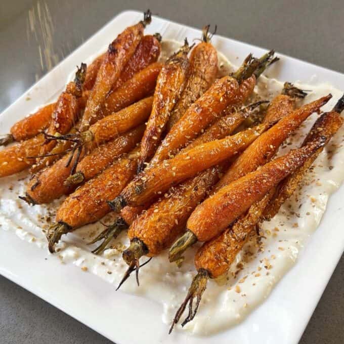 Roasted Carrot with Feta Whip Roasted whole carrots are served on a white plate over a layer of creamy white sauce and sprinkled with sesame seeds.