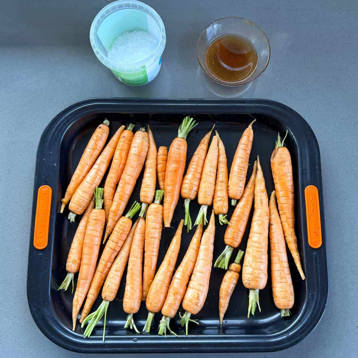 Roasted Carrot with Feta Whip A tray of fresh carrots with stems, a container of coarse salt, and a small bowl of brown liquid on a gray surface.