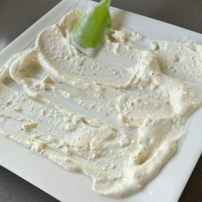Roasted Carrot with Feta Whip A green spatula spreads a thick, creamy white sauce evenly across a square white plate.