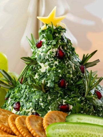 A cheese ball shaped like a Christmas tree, covered with chopped herbs, rosemary sprigs, and pomegranate seeds, topped with a cheese star, surrounded by crackers, grapes, and sliced cucumber.