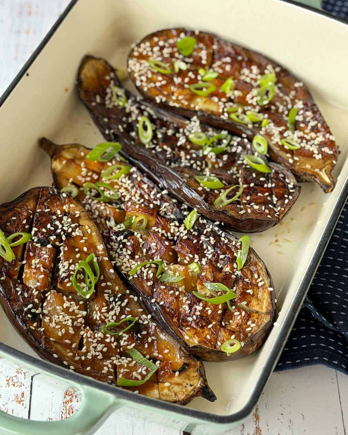 Four halved eggplants are roasted in a baking dish, topped with sesame seeds and sliced green onions.