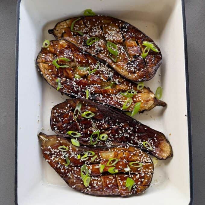 BBQ Miso Eggplant Four roasted eggplant halves are topped with sesame seeds and sliced green onions in a white baking dish.