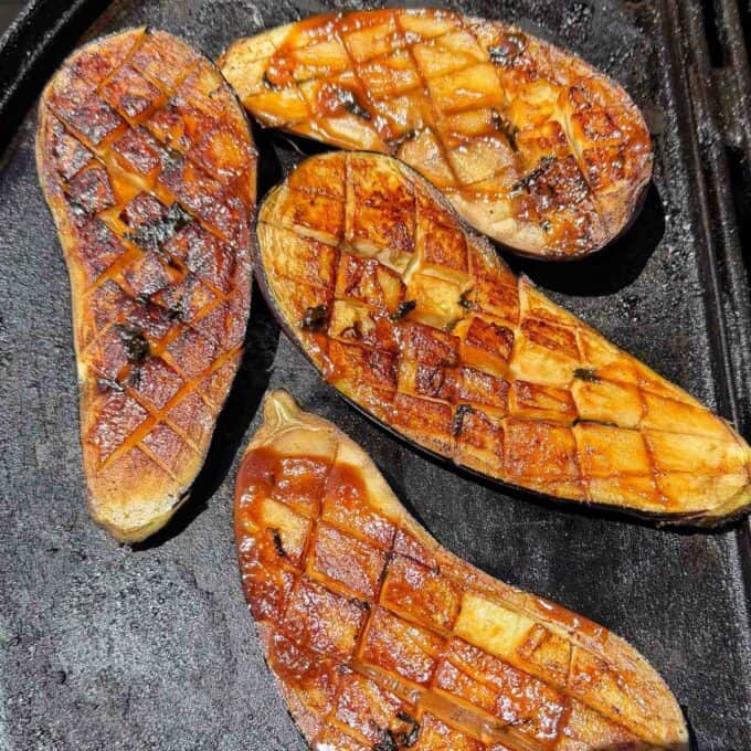BBQ Miso Eggplant Four halved eggplants with scored surfaces are grilling on a dark stovetop or grill pan, showing browned and caramelized areas.